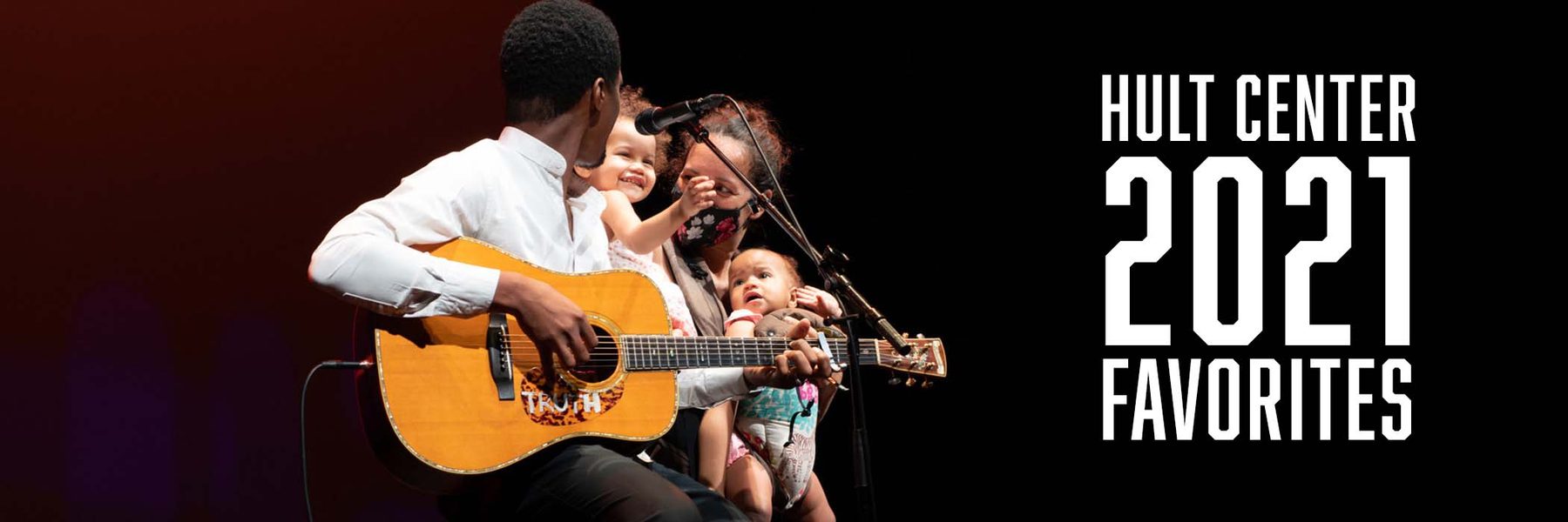 A man sitting on a stool on stage holding a guitar and a microphone in front of him with his wife and two children beside him.
