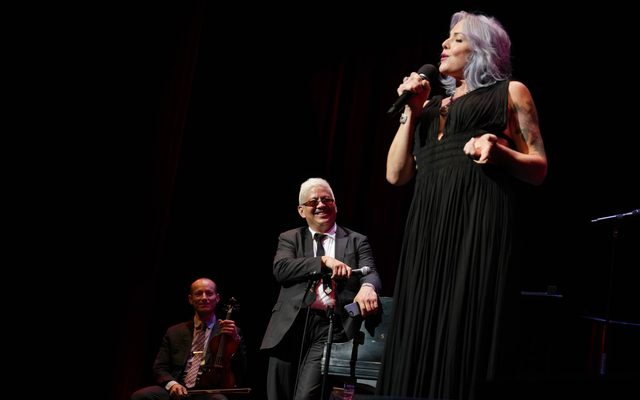 Photo of woman with brightly colored hair singing into a microphone and two musicians behind her watching