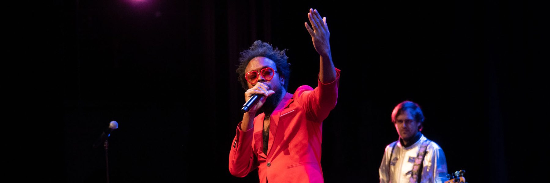 A man in a bright colored suit and heart shaped sunglasses singing into a microphone and a musician in the background.