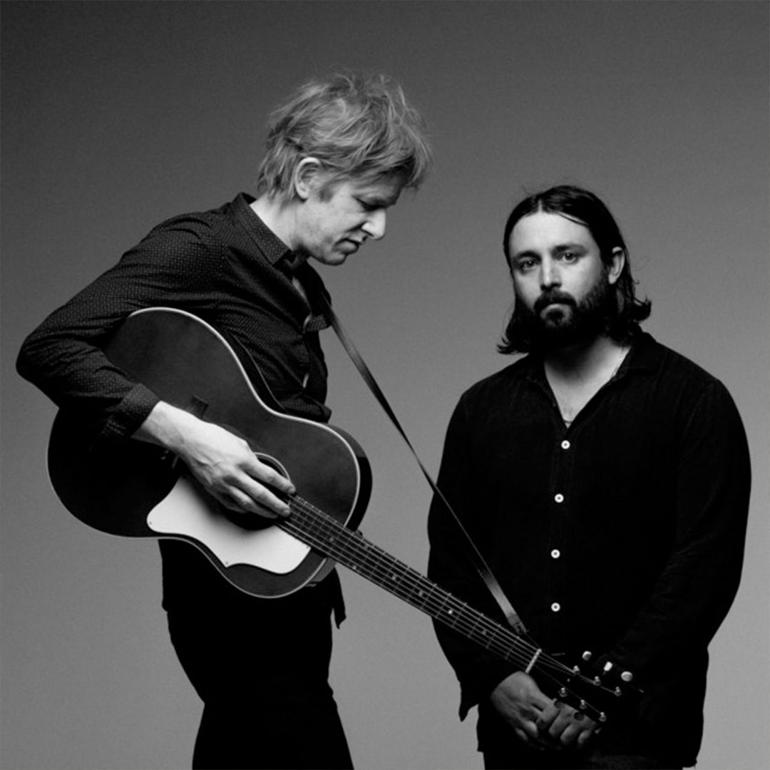 Black and white image of 2 men: taller one holding a guitar looking away from camera and shorter one looking directly at camera