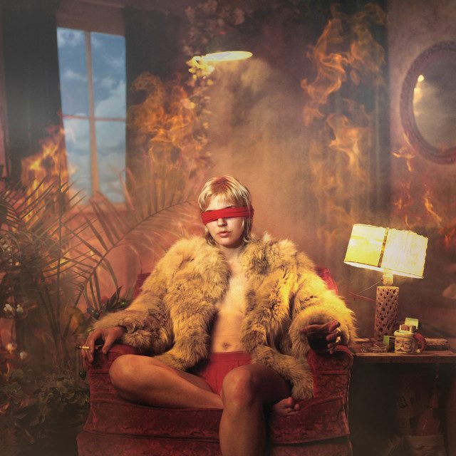 Image of a woman sitting on a sofa chair topless in fur coat and orange shorts and matching blindfold with a cigarette in their right hand and a glass with a dark liquid in it in their left hand in a room filled with smoky fire